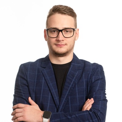 Man with short blond hair and glasses, wearing a blue plaid blazer over a black shirt, arms crossed, neutral expression.