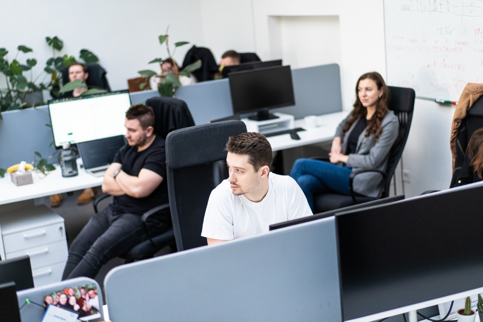 Open office scene with several employees at their desks; foreground man in a white T‑shirt looks to the right, others at computers, plants along partitions, and a whiteboard in the background.