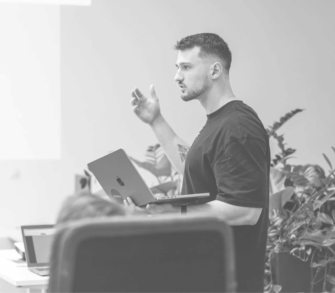Man in a black T‑shirt speaks in an office, holding a MacBook and gesturing with his right hand.