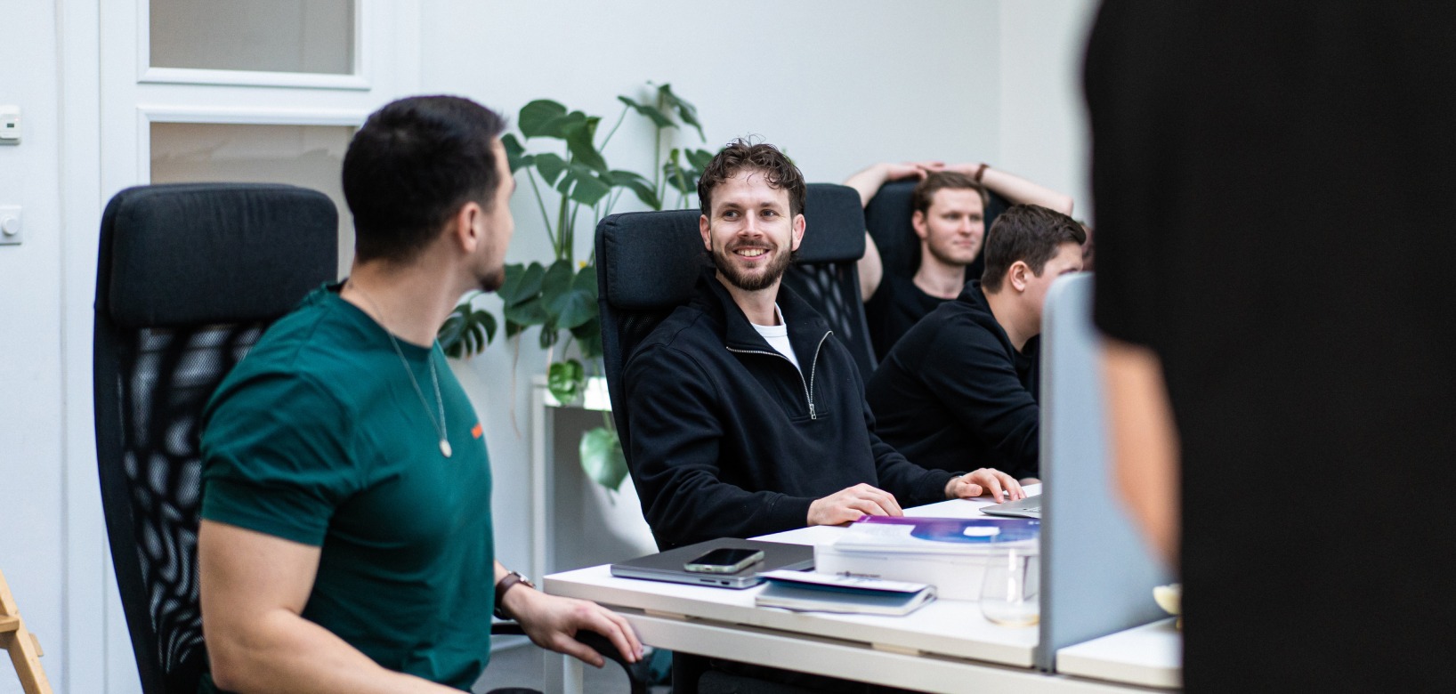 Smiling man in a black zip-up sweater sits at a white desk among coworkers in a bright office.