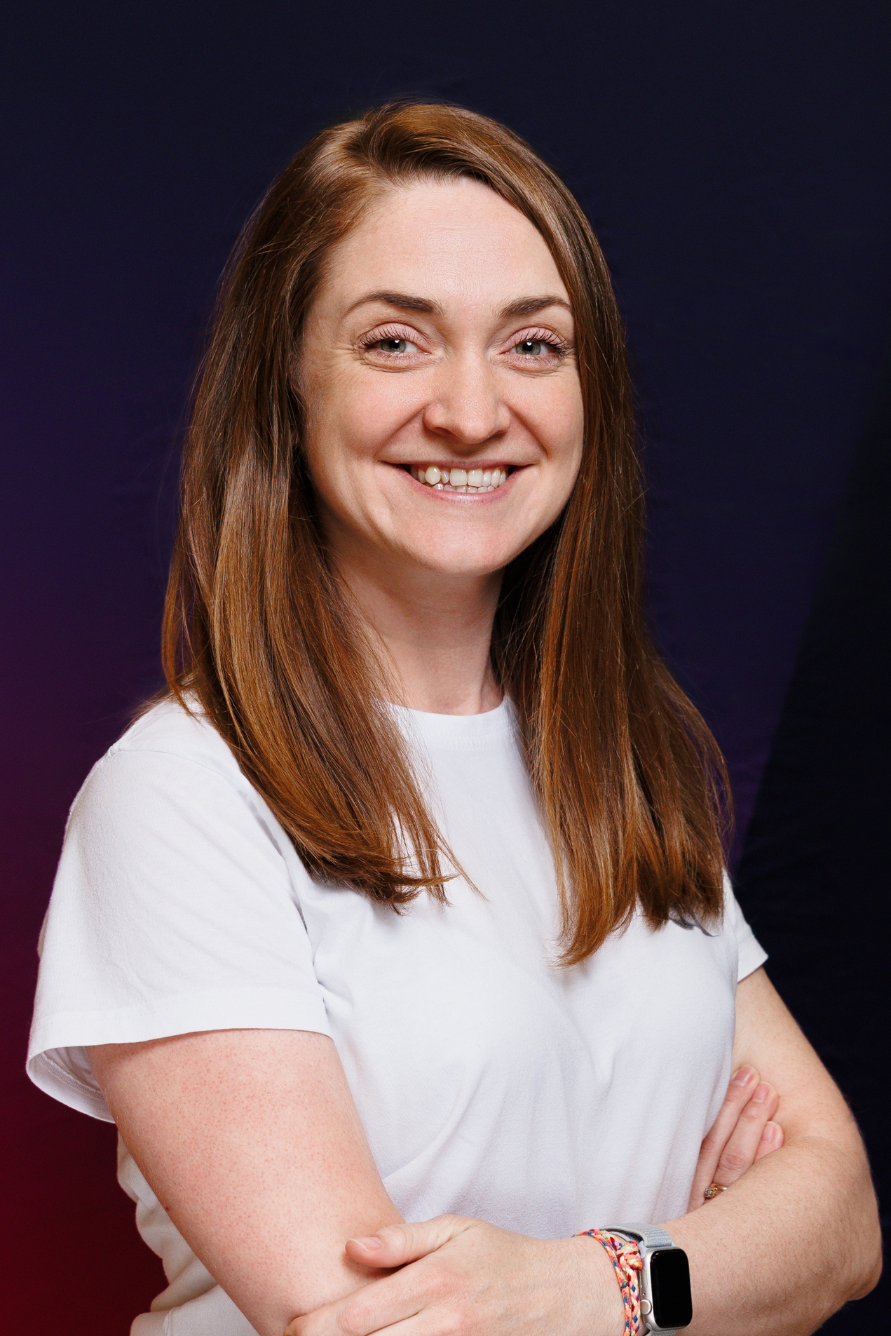 Smiling woman with long brown hair in a white shirt, arms crossed, wearing a smartwatch and colorful bracelets against a dark gradient background.